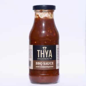 Flaske med Thya BBQ sauce, sort etiket, hvid tekst, sort låg, synlig brun sauce indeni.