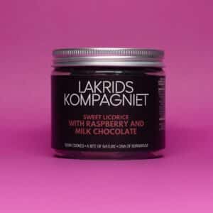 Mørk lakridsglas med metallåg på pink baggrund. Etikette: Hindbær og mælkechokolade.