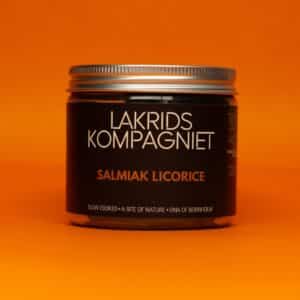 Glas med Lakrids Kompagniet Salmiak Lakrids, sort design, sølvlåg, orange baggrund.